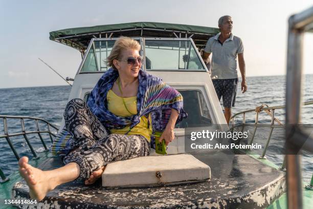una mujer positiva europea activa de 50 años, una turista, descansa en la cubierta de un pequeño barco de pesca frente a la cubierta durante el viaje de pesca en alta mar en sri lanka y está disfrutando del paseo por el océano. - 50 54 years fotografías e imágenes de stock