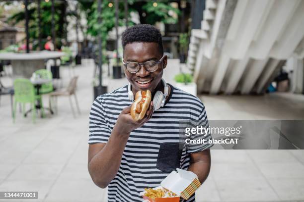 young african-american man is eating hot dog on the go - eat man stock pictures, royalty-free photos & images