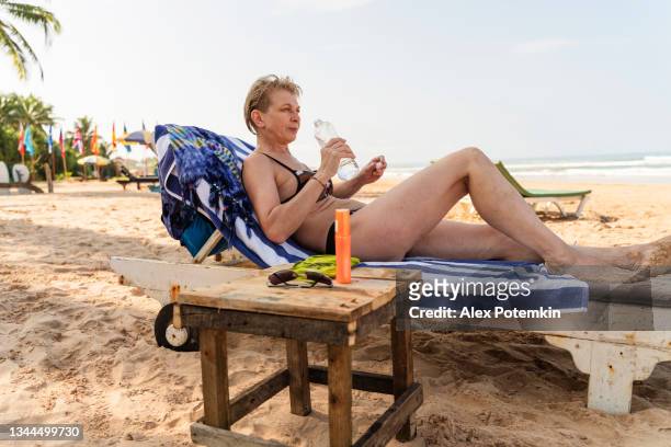 una atractiva y activa mujer europea de 50 años, turista, descansa en una tumbona en una playa de bentota, sri lanka. ella está bebiendo agua de una botella de plástico. - 50 54 years fotografías e imágenes de stock