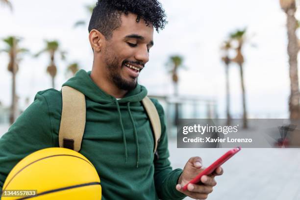 happy american man with ball using mobile phone in the street - herren basketball stock-fotos und bilder