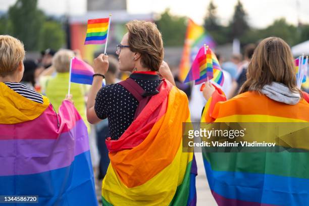 wrapped in bisexual flag and pride flags this trio are watching a gay pride event - lgbtqia rights stock pictures, royalty-free photos & images