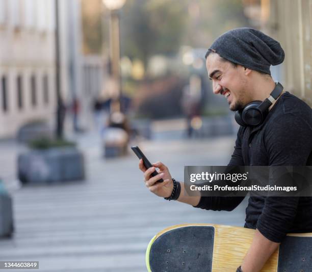 un joven viajero feliz está disfrutando de vacaciones y explorando la ciudad sosteniendo una patineta y usando un teléfono móvil. - visits music choice fotografías e imágenes de stock
