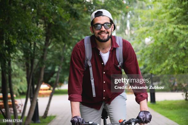 a serious young indian man, a dark-skinned indian student with glasses rides a bicycle on the street, in a public park on a bicycle path, against a background of green trees and shrubs. the concept of a healthy lifestyle, extreme sports, active recreation - holy city park bildbanksfoton och bilder