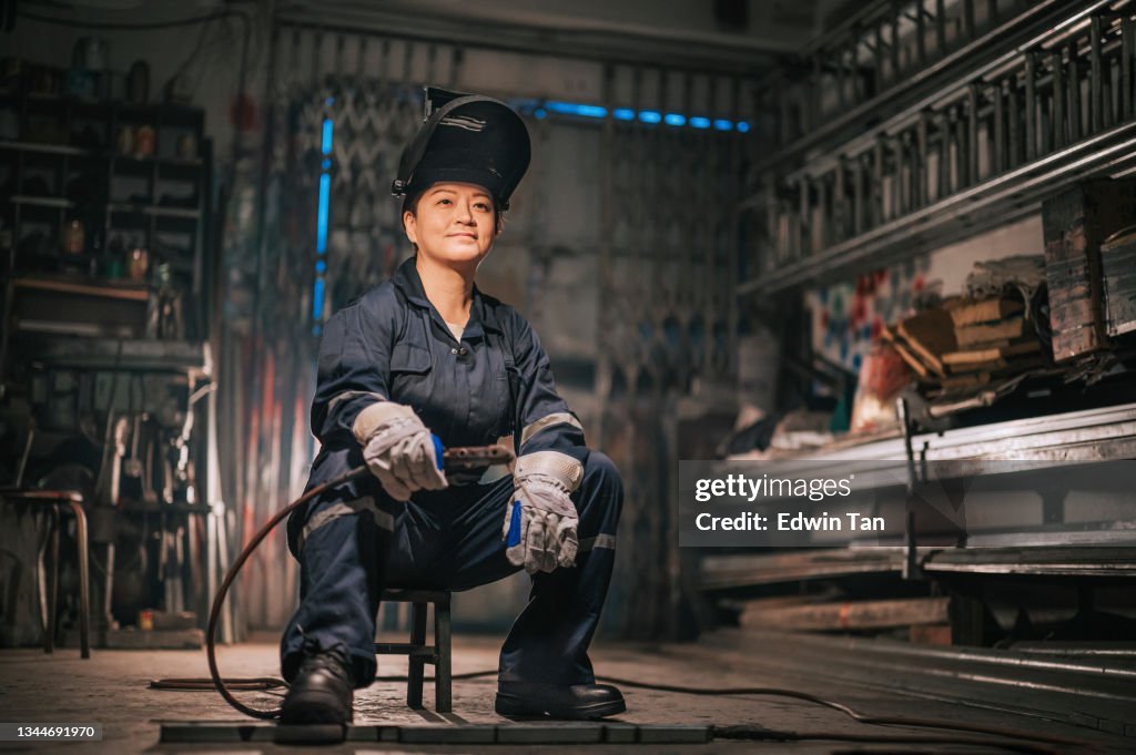 Asiatische chinesische Arbeiterin Schweißerin mit schutzschützender Arbeitskleidung, die lächelnd in der Werkstattgarage auf Hocker sitzt