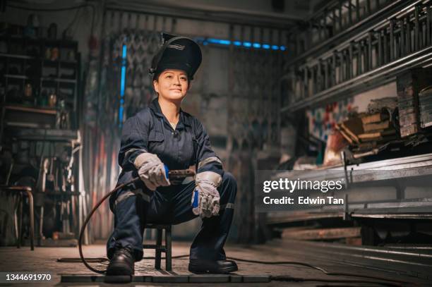 asiatica cinese donna operaia saldatrice con indumenti da lavoro protettivi che distoglie lo sorridino nel garage dell'officina seduta sullo sgabello - tough lady foto e immagini stock