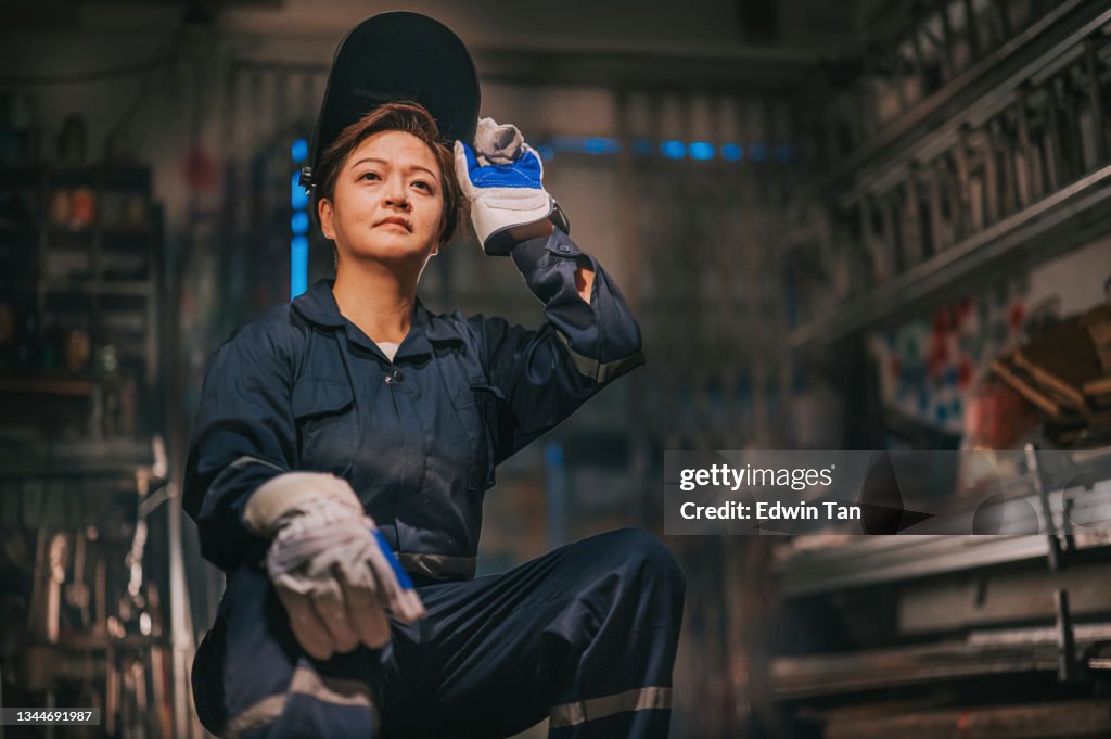Asian chinese female blue collar worker welder with protective workwear looking away smiling in workshop garage sitting on stool