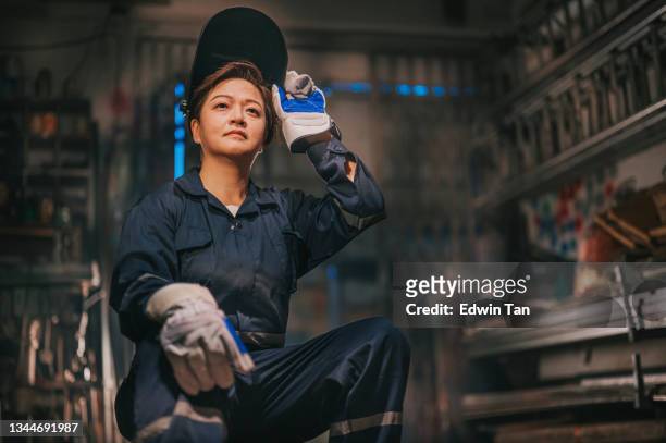 asian chinese female blue collar worker welder with protective workwear looking away smiling in workshop garage sitting on stool - welding mask stock pictures, royalty-free photos & images