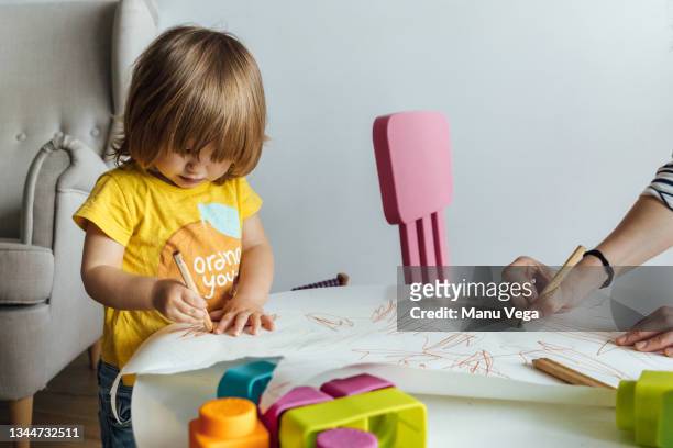 cropped unrecognizable adult hand helping cute little girl in yellow shirt sitting at table drawing with colorful pencils while spending time in playroom in kindergarten - babysitter stock pictures, royalty-free photos & images