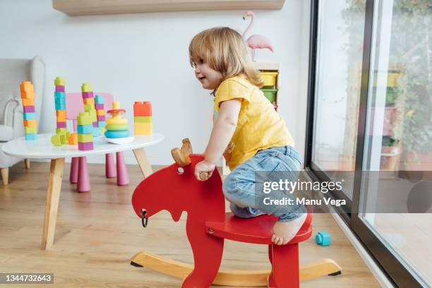 side view of cute infant child in casual clothes having fun and riding red wooden horse while playing in kindergarten - balançoire à bascule photos et images de collection