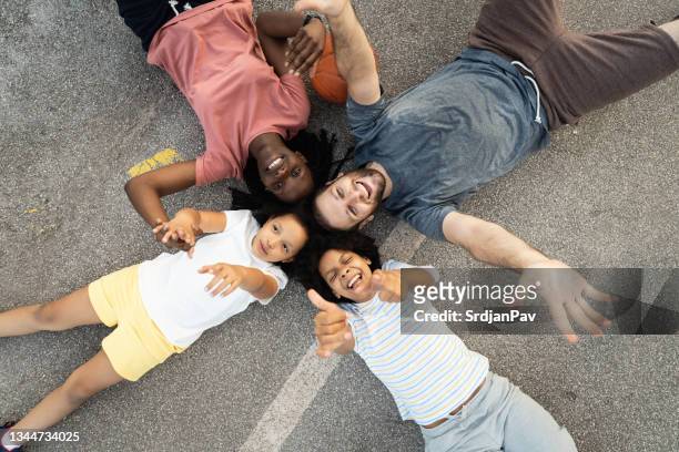 happy multiracial stepfamily, lying on the sports basketball court and relaxing after a basketball game - blended family stockfoto's en -beelden