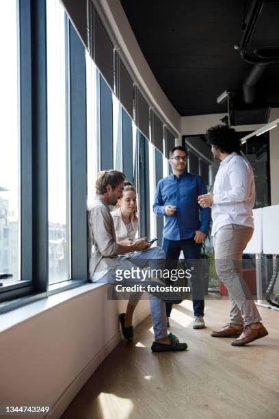 quatre collègues d’affaires qui traînent près de la fenêtre du bureau pendant la pause-café - pause café bureau photos et images de collection