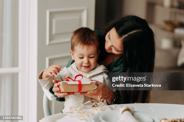 birthday celebration concept. happy mom gives a surprise gift box to her little son child sitting hugging at the dinner table on christmas day in a cozy room at home in winter indoor - child giving gift stock-fotos und bilder