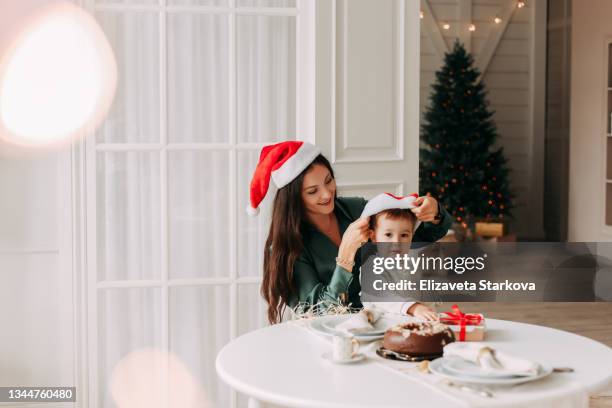 the concept of christmas. happy cheerful young mother and little son child in red santa claus hats are sitting at a festive dining table celebrating winter holidays, mom congratulates gives a gift box to the kid in a decorated room with a christmas tree - happy holidays family stock pictures, royalty-free photos & images