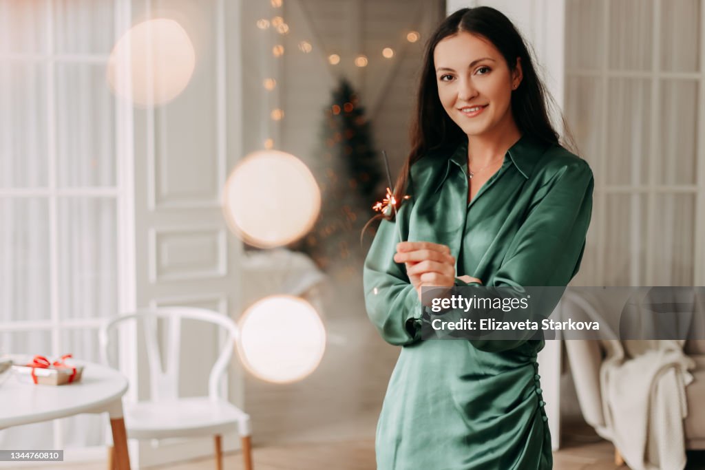 A happy beautiful young woman in an elegant emerald dress smiles burns a sparkler celebrates the Christmas holiday on a winter weekend in a cozy room of the indoor house