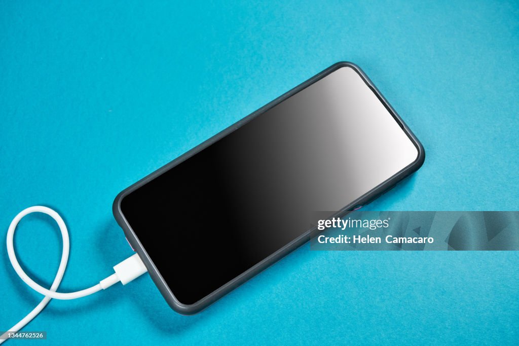 Mobile phone connected to electricity for recharging on blue background.