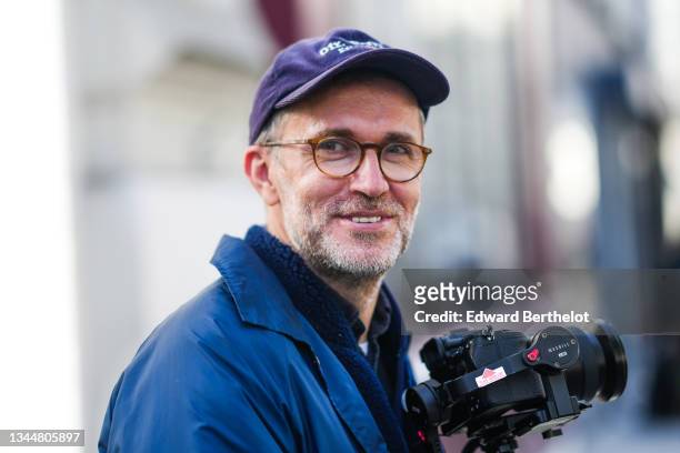 Loic Prigent wears a navy blue ribbed velvet cap with white embroidered inscriptions, glasses, a navy blue fluffy high-neck zipper pullover form...