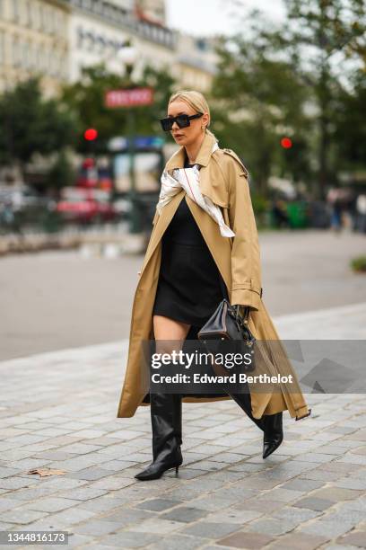 Justyna Czerniak wears black sunglasses, gold earrings, a beige long oversized trench coat, a white with red and gray print pattern silk scarf, a...