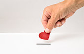 Red heart symbol is put by person's hand into slot of white donation box
