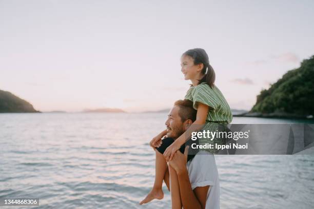 father carrying young daughter on shoulders on beach at sunset - family traveling stock-fotos und bilder