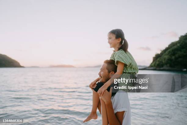 father carrying young daughter on shoulders on beach at sunset - vacaciones de sol y playa fotografías e imágenes de stock