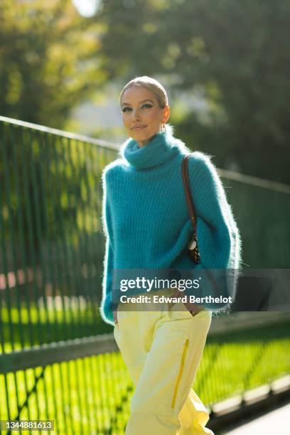 Leonie Hanne wears a gold and silver large pendant earrings, a blue fluffy turtleneck oversized pullover, a brown shiny leather with a chain shoulder...