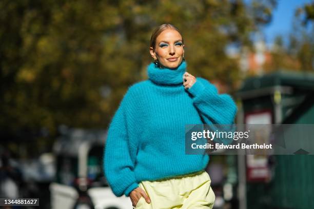 Leonie Hanne wears a gold and silver large pendant earrings, a blue fluffy turtleneck oversized pullover, a brown shiny leather with a chain shoulder...