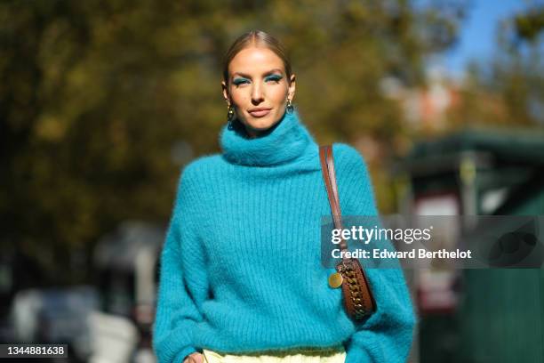 Leonie Hanne wears a gold and silver large pendant earrings, a blue fluffy turtleneck oversized pullover, a brown shiny leather with a chain shoulder...