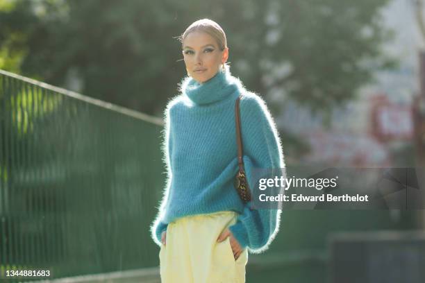 Leonie Hanne wears a gold and silver large pendant earrings, a blue fluffy turtleneck oversized pullover, a brown shiny leather with a chain shoulder...