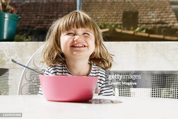 adorable little child sitting at table with bowl on terrace and smiling during breakfast in morning with eyes closed - child eyes closed stock pictures, royalty-free photos & images