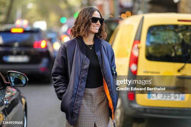 Erika Boldrin wears black sunglasses, a silver chain pendant necklace, a black t-shirt, a navy blue oversized bomber coat, a high waist beige with...