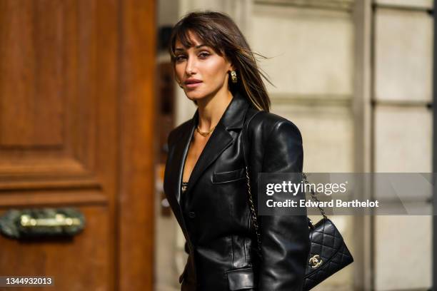 Guest wears gold large earrings, a gold necklace, a black shiny leather blazer dress with shoulder pads, a black V-neck t-shirt, a black shiny...