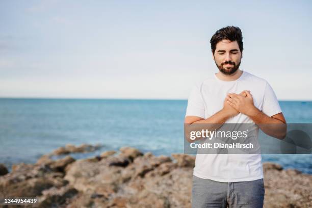 handsome young man with hands on chest, closed eyes and grateful face gesture in front of the sea - self love stock pictures, royalty-free photos & images