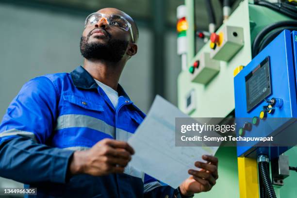 african american engineer hand hold machine manual book paper checking check list for safty process manufacturing system,smart technician double checking peper report for daily cnc press machine operation in technilogy factory - macacão preto imagens e fotografias de stock