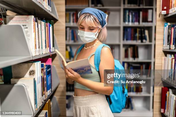 young girl wearing protective face mask choosing book from library - library mask stock pictures, royalty-free photos & images