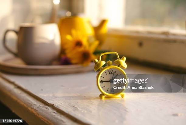 fall seasonal still life. retro alarm clock. fall back concept. cup with autumn flowers and kettle on wooden tray on window sill - daylight saving time foto e immagini stock