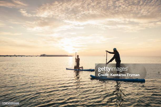 man learning to paddleboard from male instructor in sea during sunset - paddelbrett stock-fotos und bilder