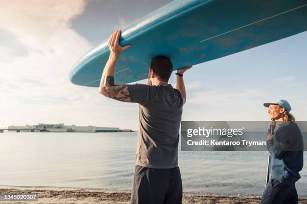man carrying paddleboard on head while male friend standing at beach - beach hold surfboard stock-fotos und bilder