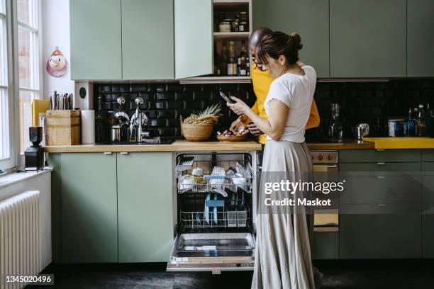 smiling woman sharing smart phone to boyfriend doing dishes in kitchen at home - dishwasher stock pictures, royalty-free photos & images