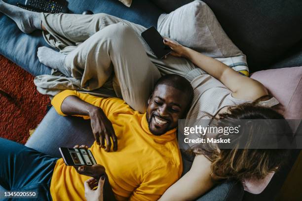 cheerful man lying on sofa with girlfriend sitting in living room at home - liebespaar stock-fotos und bilder