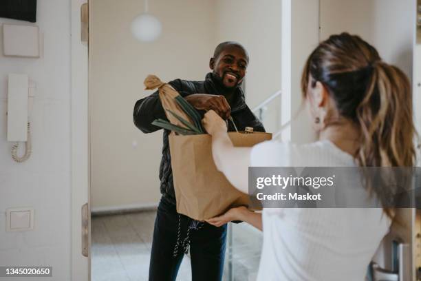 smiling man giving paper bag of groceries to woman standing at doorway - delivery stock-fotos und bilder