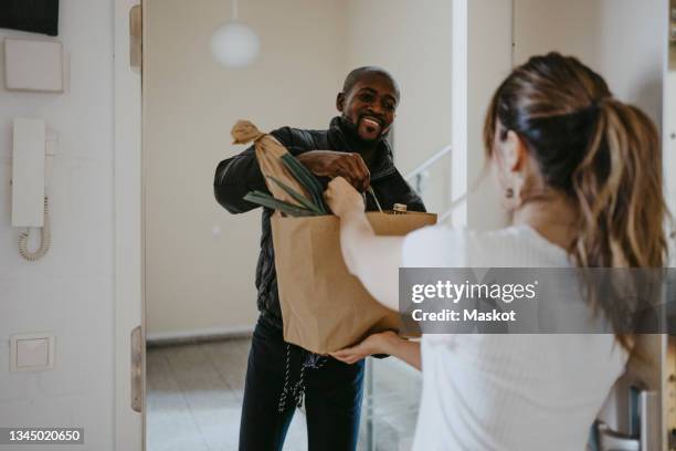 smiling man giving paper bag of groceries to woman standing at doorway - consegna a domicilio foto e immagini stock
