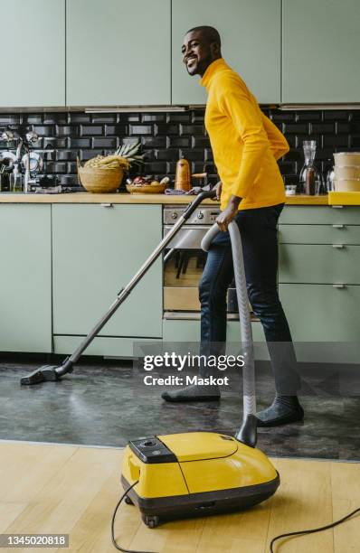 full length of smiling man using vacuum cleaner doing chores in kitchen at home - saugen stock-fotos und bilder