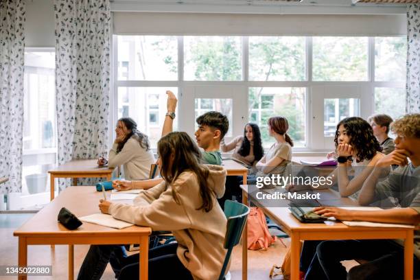 boy raising hand while sitting with other students in classroom - highschool stock pictures, royalty-free photos & images