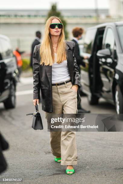 Pernille Teisbaek wears green sunglasses, earrings, gold chain necklace, a white ribbed V-neck t-shirt, a black shiny leather long blazer jacket, a...