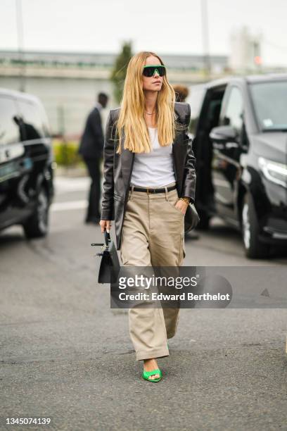 Pernille Teisbaek wears green sunglasses, earrings, gold chain necklace, a white ribbed V-neck t-shirt, a black shiny leather long blazer jacket, a...