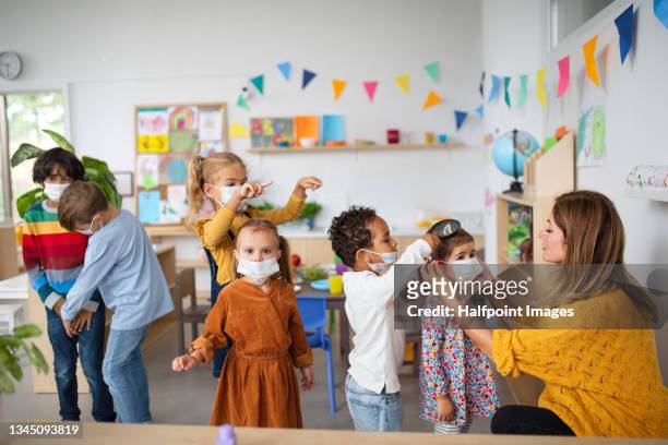 pre school teacher helping little girl to wear face mask indoors at nursery school, coronavirus concept. - child coronavirus sick stock pictures, royalty-free photos & images
