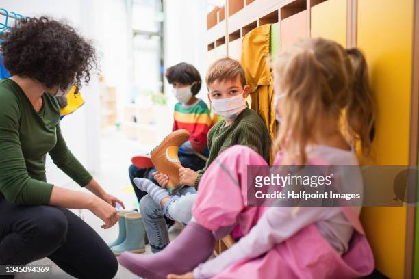 pre school teacher helping children to put on shoes indoors in cloakroom at nursery, coronavirus concept. - child coronavirus sick stock pictures, royalty-free photos & images