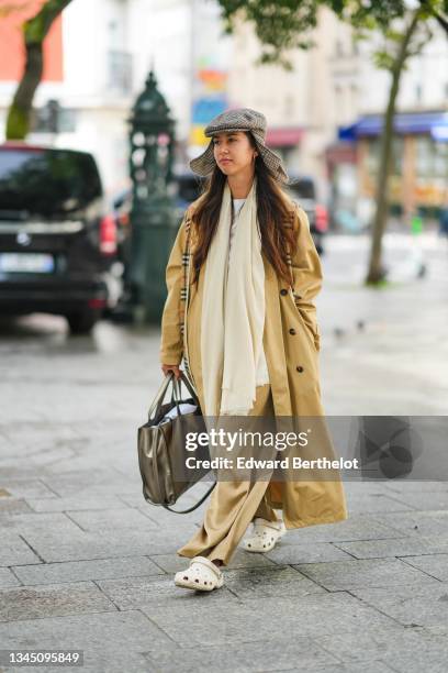 Guest wears a gray and burgundy checkered pattern wool beret with long ear, gold earrings, a beige long oversized trench coat, a beige with red and...