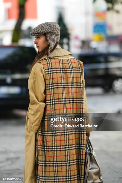 Guest wears a gray and burgundy checkered pattern wool beret with long ear, gold earrings, a beige long oversized trench coat, a beige with red and...