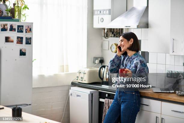 cheerful woman on phone in kitchen drinking coffee - looking at phone stock-fotos und bilder